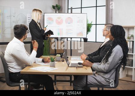 Schöne motivierte, smarte, blonde junge Geschäftsfrau, die vor ihrem konzentrierten multiethnischen Geschäftsteam Infografiken auf dem digitalen Brett vorführt, ihr zuhört und Notizen macht. Stockfoto