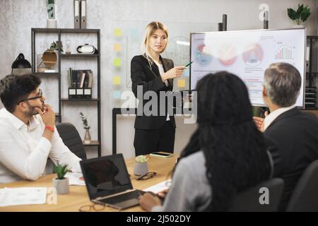 Schöne motivierte, smarte, blonde junge Geschäftsfrau, die vor ihrem konzentrierten multiethnischen Geschäftsteam Infografiken auf dem digitalen Brett vorführt, ihr zuhört und Notizen macht. Stockfoto