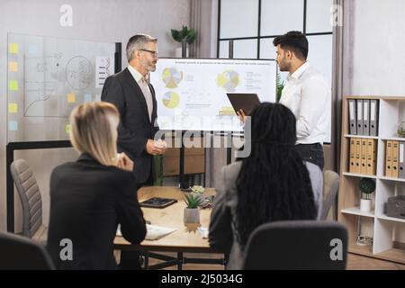 Ein Team aus verschiedenen aufmerksamen Geschäftsleuten verschiedener Rassen, die ihrem männlichen bärtigen Chef und indischen Manager mittleren Alters zuhören, zeigt die Dynamik des wichtigsten Weltmarktes mithilfe eines digitalen Wandbildschirms. Stockfoto