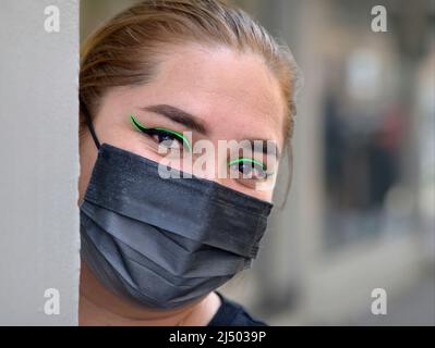 Junge schöne mexikanische Latina Frau mit markantem grünen Eyeliner trägt schwarze chirurgische Gesichtsmaske und schaut um eine Ecke auf den Betrachter. Stockfoto