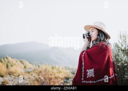 Eine junge Frau mit Sommerstimmung, die einen roten Poncho und einen Hut trägt und mit einer Vintage-Kamera Fotos in einem natürlichen Raum macht. Stockfoto