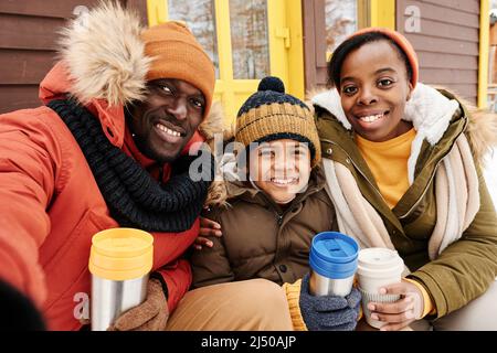 Junge, fröhliche afroamerikanische Familie mit drei Kindern, die bei ihrem Landhaus einen heißen Tee trinken und die Kamera betrachten, während sie ein Selfie-Portrait machen Stockfoto