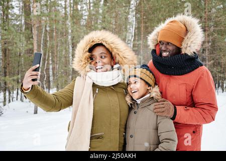Glückliche, liebevolle afroamerikanische Familie in warmer Winterkleidung, die Selfie im Park oder Wald macht, während die Frau das Smartphone hält Stockfoto