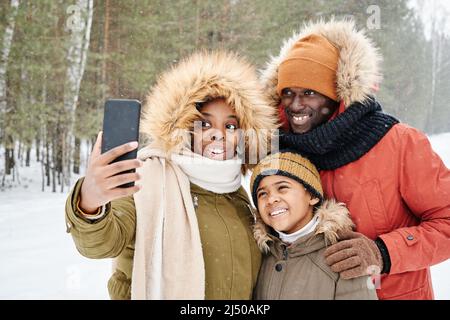 Glückliche junge Familie von Eltern und ihren niedlichen Sohn in warmen Winterjacken machen Selfie im Freien, während Freizeit in Park oder Wald Stockfoto
