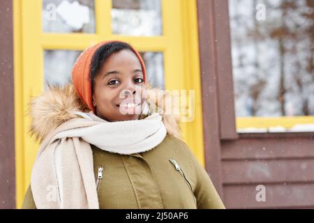 Fröhliche afroamerikanische Frau in Winterjacke, Mütze und weißem Wollschal, die vor der Kamera gegen das neue Landhaus schaut Stockfoto