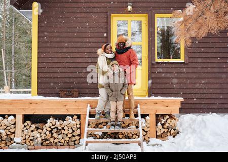 Drei glückliche afroamerikanische Familienmitglieder stehen vor der Veranda des neuen Landhauses, während sie den verschneiten Wintertag genießen Stockfoto