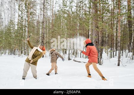 Junge afroamerikanische Frau in Winterkleidung, die am Wintertag Selfie mit ihrem Sohn und Ehemann macht und Schneebälle im Wald spielt Stockfoto