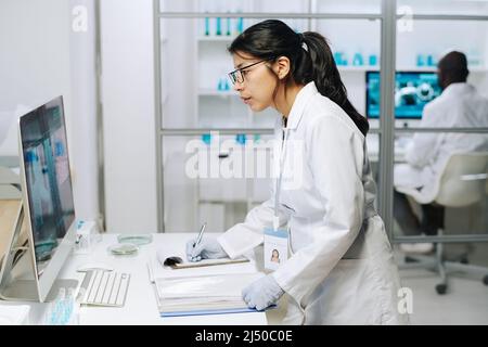 Seitenansicht einer jungen, seriös klinikenden Ärztin, die den Computerbildschirm mit einem Coronavirus-Makrobild betrachtet und dabei Notizen im Dokument macht Stockfoto