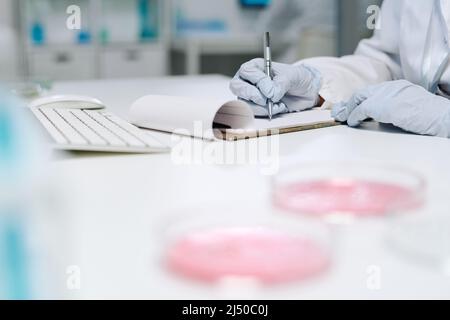 Handschuhen eines modernen Klinikers oder Biochemikers, der die Ergebnisse wissenschaftlicher Experimente am Arbeitsplatz aufschreibt Stockfoto