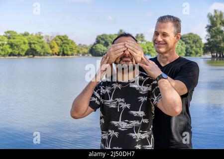 Ein paar ältere schwule Männer, einer überraschte den anderen, indem er seine Augen in einem See verhüllte. Konzept der Überraschung, Glück, Spiel Stockfoto