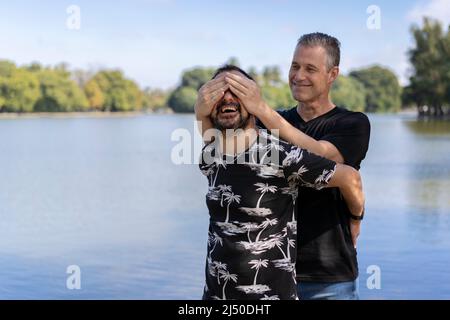 Ein paar ältere schwule Männer, einer überraschte den anderen, indem er seine Augen in einem See verhüllte. Konzept der Überraschung, Glück, Spiel Stockfoto