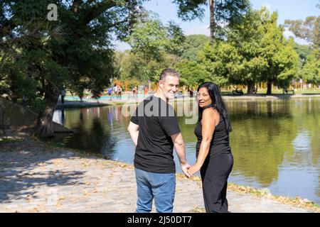Multiethnisches Paar, das aus einer Andenfrau und einem kaukasischen Mann besteht, der an einem See entlang geht. Fröhliche Ausdrücke und Gesichter von Liebenden Stockfoto
