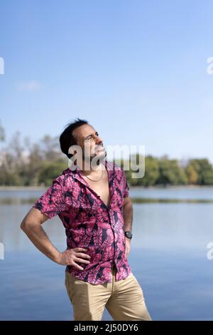 Bärtiger reifer Mann in einem fuchsienfarbenen Hemd, Arme Akimbo an einem See Stockfoto