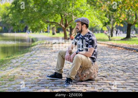 Reifer Mann sitzt auf einem Baumstamm und schaut pensiver auf einen See. Speicherplatz kopieren Stockfoto