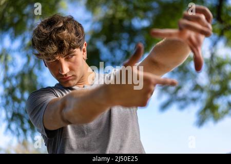 Ein hübscher junger Mann blinzelt die Augen und schaut durch einen Rahmen aus Händen. Attraktiver Mann machen Bild für Finger, isoliert auf unscharfen Hintergrund Witz Stockfoto