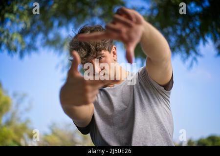 Ein hübscher junger Mann blinzelt die Augen und schaut durch einen Rahmen aus Händen. Attraktiver Mann machen Bild für Finger, isoliert auf unscharfen Hintergrund Witz Stockfoto