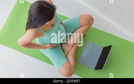 Schwangere beobachten pränatalen Online-Kurs zu Hause auf Laptop. Draufsicht auf die Fitnessmatte. Schwangerschaft Konzept des Mädchens üben Atemtechniken Stockfoto
