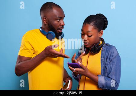 Schockierte Männer und Frauen, die auf den Bildschirm des Mobiltelefons zeigen und unerwartete Nachrichten in den sozialen Medien finden. Überrascht Paar Spaß mit Kopfhörern und modernen Smartphone im Internet zu surfen. Stockfoto