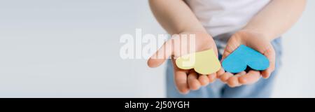 Teilansicht des Kindes mit blauen und gelben Papierherzen auf offenen Handflächen isoliert auf grauem Banner Stockfoto