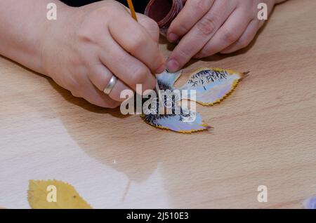 Die Hand einer erwachsenen Frau zieht eine Eule auf ein trockenes Baumblatt. Bunte Zeichnung eines Vogels auf einem gefallenen Blatt. Zeitgenössische Kunst Stockfoto