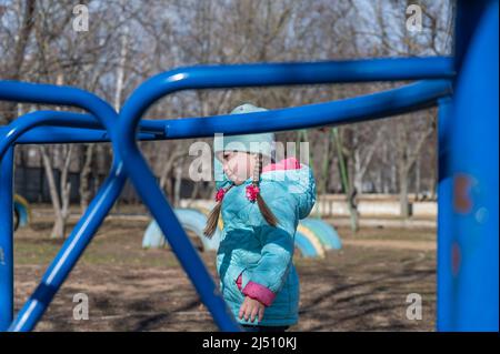 Auf dem Spielplatz steht ein Mädchen mit einem langen Zopf. Ein fünfjähriges Kind ohne Pflege in der Welt. Blaue Kinderschaukel im Vordergrund. Stockfoto