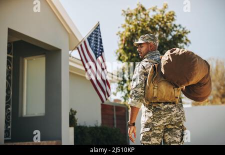 Mutiger junger Soldat, der mit seinem Gepäck auf sein Haus zugeht. Rückansicht eines patriotischen amerikanischen Militäranmannes, der nach seinem Dienst nach Hause zurückkehrt Stockfoto