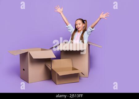 Foto von entzückend beeindruckt kleine Schule Mädchen tragen Denim Hemd sitzen in großen Kasten steigende Arme isoliert lila Farbe Hintergrund Stockfoto
