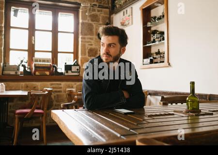 Nachdenklicher Mann, der an einem alten Ort auf seinen Kaffee wartet, der bärtige Mann in einer Denim-Jacke hat seine Arme gefaltet und sein Gesicht zeigt, dass er gelangweilt ist, t Stockfoto