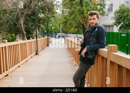 Der junge bärtige, gutaussehende Mann posiert, trägt eine Denim-Jacke, lehnt sich auf die Brücke und posiert für die Kamera, hat eine ernsthafte Gesichtsausdruck Stockfoto