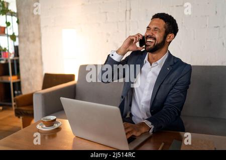 Fröhlicher junger Geschäftsmann, der auf dem Sofa sitzt und im Büro am Handy telefoniert. Stockfoto