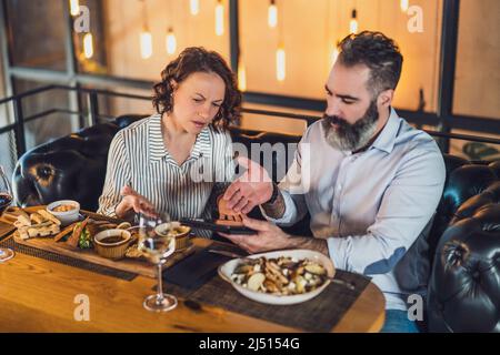 Das Paar sitzt im Restaurant. Sie essen zu Mittag, nutzen digitale Tablets und streiten sich. Stockfoto