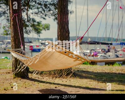 boho-Stil weiße Leinenhängematte, die zwischen Pinien an einem Seeufer mit Segelbooten Hafen im Hintergrund hängt Stockfoto