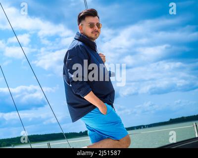 Der junge Mann mit schwarzen Haaren trägt eine Sonnenbrille und ein nautisches Tuch und steht während seiner Segelreise im Sommer auf einem Segelboot Stockfoto