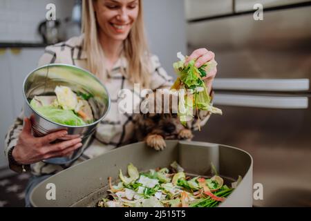 Frau wirft Gemüseschnitzel in einen Komposteimer in der Küche und füttert Hund. Stockfoto