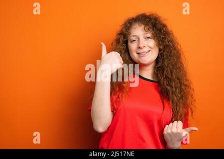 Attraktive Rothaarige Fußballsportfan Frau trägt Mannschaftsuniform und hält Daumen nach oben auf orangefarbenem Hintergrund. Stockfoto