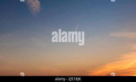 Passagierflugzeug, das bei Sonnenuntergang am blauen Himmel fliegt, lässt einen schnellen Transport hinter einem weißen Kondensstreifen zurück. Eine Spur von Kondensstreifen oder Dampfspuren, Stockfoto