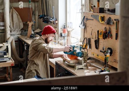 Der hohe Winkel des jungen kaukasischen Zimmermanns, der in der Werkstatt ein Stück des Holzbodens mit der Gehrungsäge schneidet Stockfoto