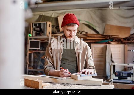 Professioneller junger Zimmermann mit Lineal und Bleistift, der in der Holzwerkstatt Markierungen auf einem Stück Holzbrett macht Stockfoto