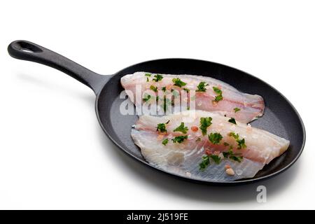 Zwei Filets aus weißem Fisch, mit Salz und gehackter Petersilie in einer gusseisernen Pfanne verschüttet. Bereit zum Braten. Isoliert auf weißem Hintergrund Stockfoto