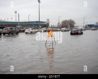 Eine überflutete Stadtstraße. Die Straße unter Wasser. Stürme und heftige Regenfälle trafen viele Teile von Tatarstan und Russland. Flut auf der Straße planschen mit einem Auto. Straßenbauschild Stockfoto