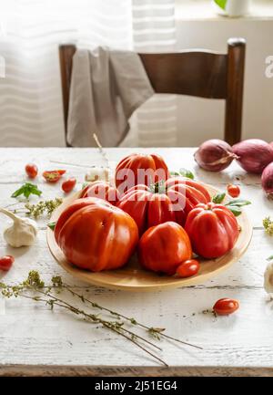 Rote Tomaten auf dem Teller und weißer Holzhintergrund mit Orangano, Basilikumblättern, Knoblauch und Zwiebeln. Stockfoto