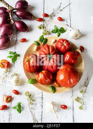 Tomaten auf dem Teller und weißer Holzhintergrund mit Orangano, Basilikumblättern, Knoblauch und Zwiebeln Stockfoto