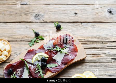 Jerky Fleisch bresaola mit Zitronen, Oliven, Grana und Rucola auf dem hölzernen Hintergrund mit Kopierraum Stockfoto