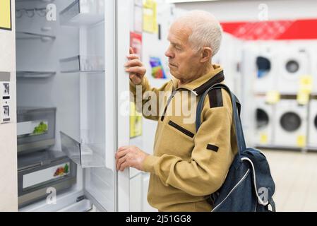 Älterer grauhaariger Rentner, der am Schalter im Ausstellungsraum der Abteilung für Elektrogeräte einen Kühlschrank sah Stockfoto