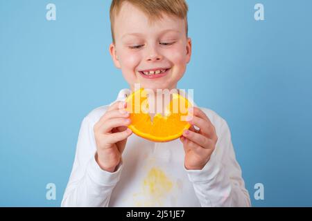 Junge hält eine geschnittene Hälfte einer Orange. Schmutzige Flecken Orangensaft auf der Kleidung. Das Konzept der Reinigung Flecken auf Kleidung Stockfoto
