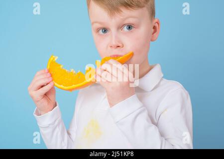 Junge essen einen Schnitt Hälfte einer Orange. Schmutzige Flecken von Orangensaft auf der Kleidung. Tägliches Leben Fleck Konzept Stockfoto