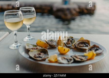 Frisch gefangenen Austern auf einem Teller und Weingläsern. Restaurant am Ufer der Bucht von Kotor in der Nähe der Austernfarm, Montenegro. Meeresfrüchte. Wunderschön Stockfoto