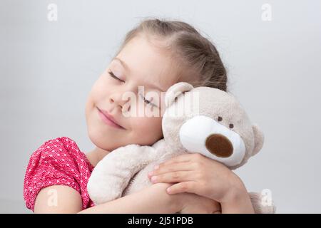 Glückliches Kind umarmt Teddybär auf weißem Hintergrund kaukasischen kleinen Mädchen von 5-6 Jahren in rot mit geschlossenen Augen Stockfoto