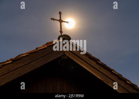 Berschis, Schweiz, 13. April 2022 der Mond steigt nachts hinter einem heiligen Kreuz einer winzigen Kapelle auf einem Hügel auf Stockfoto