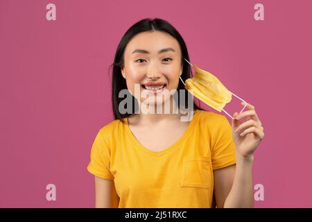 Ende der Coronavirus-Pandemie. Glückliche asiatische Dame, die medizinische Maske auszieht und sich über violetten Studiohintergrund frei fühlt Stockfoto